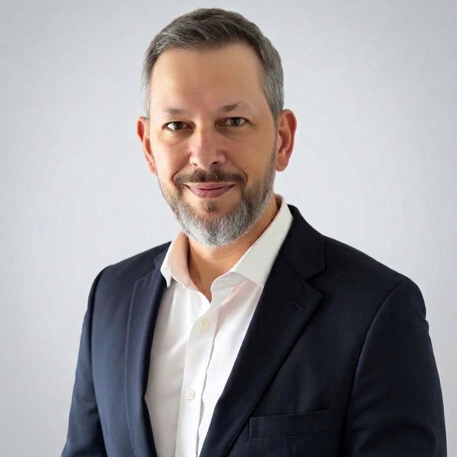 Alan Prachar wearing a navy blazer and white shirt against a light neutral background.